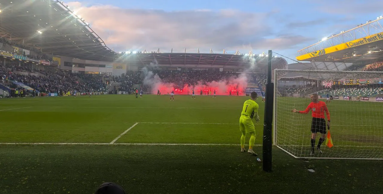 Pitch-side view of a NIFL match at a packed stadium, with red flares in the stand behind the goal